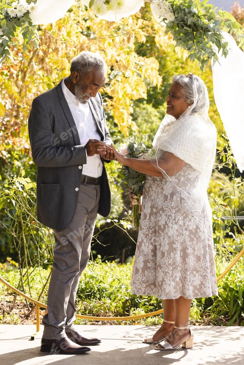 Preview: Happy senior biracial groom putting ring on finger of bride at sunny outdoor wedding ceremony
