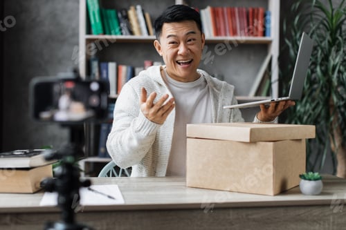 Preview: Asian man recording video on phone camera while unpacking box with new wireless laptop
