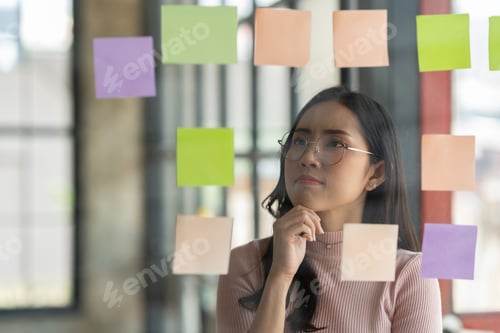 Preview: Confident businesswoman wearing eyeglasses looking and thinking Idea goes on post-it note on glass w