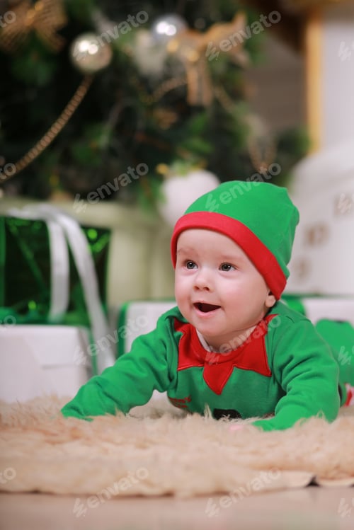 Preview: baby boy in red green elf costume sitting under Christmas tree and gift boxes