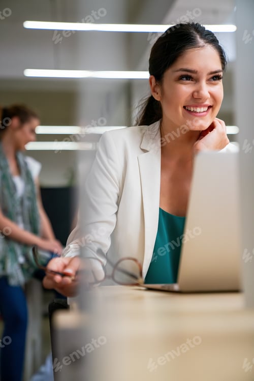 Preview: Successful young businesswoman working on laptop in office with colleagues at the background.