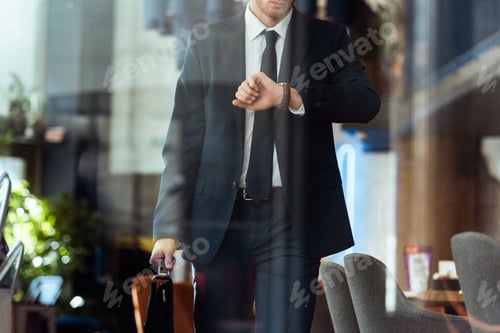 Preview: partial view of businessman with suitcase checking time while walking in cafe
