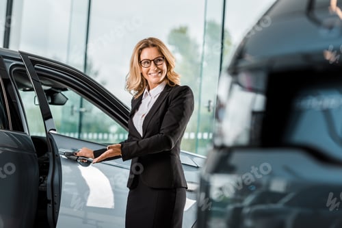 Preview: attractive smiling female car dealer opening car in showroom