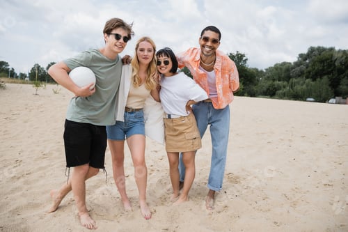 Preview: happy barefoot multiethnic friends with ball looking at camera on beach
