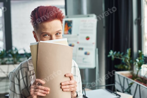 Preview: cheerful bigender person with short hair obscuring face with folders and looking at camera in office