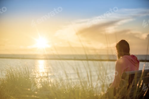 Preview: Female sitting on a bench near the sea with the sun shining in the blurred background
