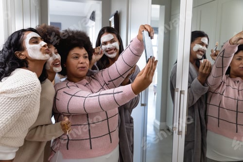 Preview: Happy biracial woman taking selfie over cellphone with female friends applied beauty cream on faces