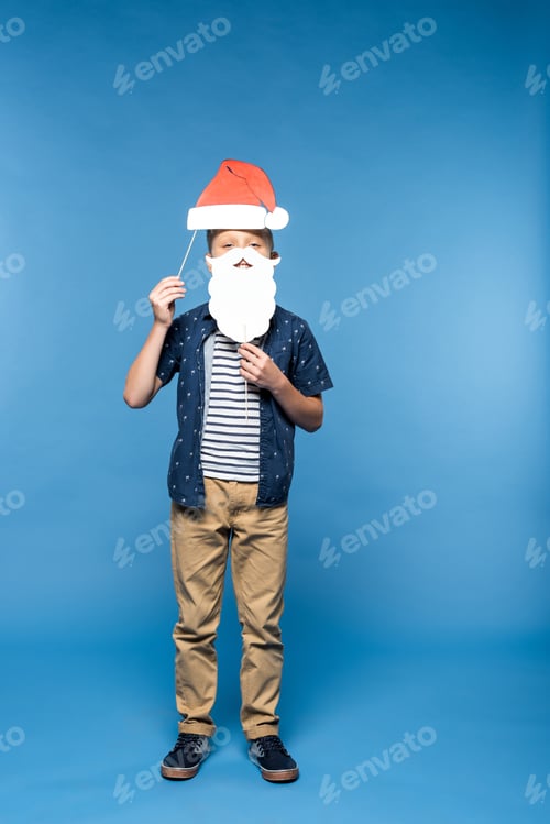 Preview: cute little boy holding paper santa hat and fake beard isolated on blue