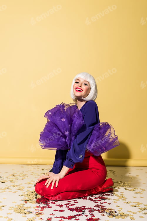 Preview: Smiling stunning girl in wig sitting on confetti on yellow background