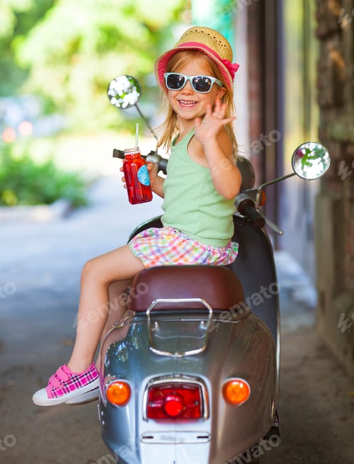 Preview: Happy little girl sitting in a vintage scooter in the street wearing hat and sunglasses