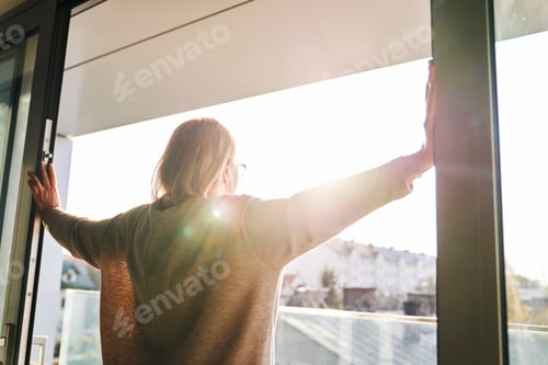 Preview: Back view of senior woman opening balcony doors in the beams of the sun