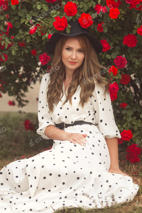 Preview: Beautiful redhead woman in hat and long dress sitting on grass and smells aroma of red rose flowers