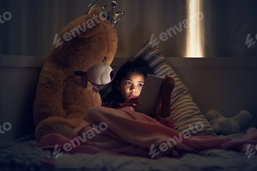 Preview: Shot of a little girl using a digital tablet while lying in bed with her teddy at night