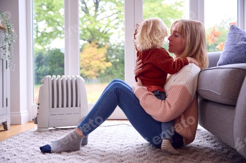 Preview: Mother With Son Trying To Keep Warm By Radiator At Home During Cost Of Living Energy Crisis