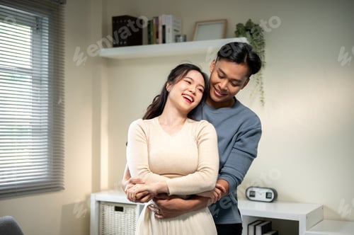 Preview: Sweet couple smiling asian man and a woman holding hands and standing hugging in living room at home