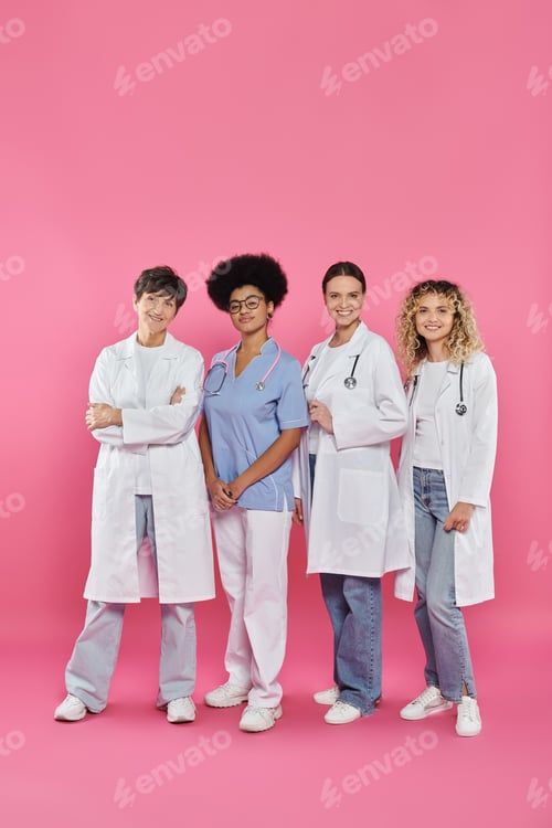 Preview: full length of smiling interracial doctors standing together on pink, breast cancer month concept