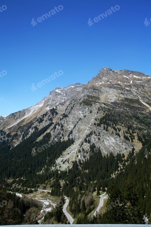 Preview: View of a winding mountain road under a clear blue sky, surrounded by green forests and rocky peaks.