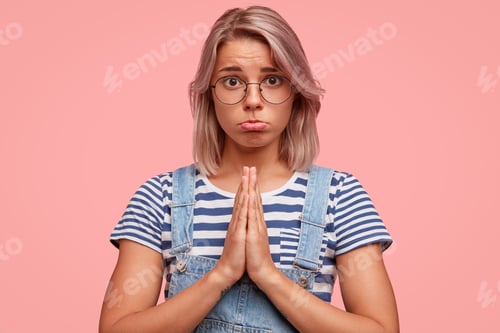 Preview: Portrait of lovely European female with sorrowful expression, makes praying gesture, curves lower li