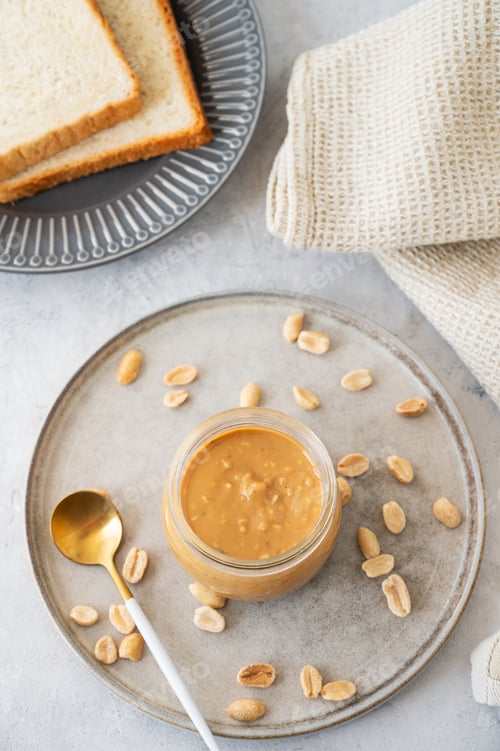 Preview: Peanut butter in glass jar on light background with salted nuts and bread toasts.