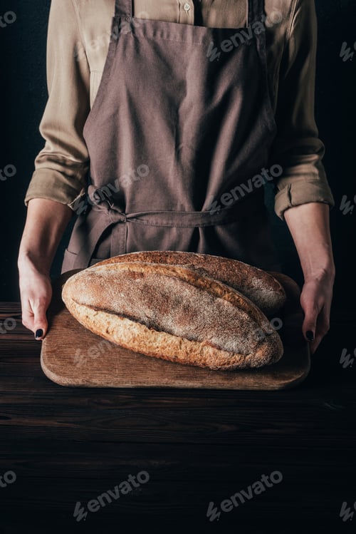Preview: partial view of woman holding loafs of bread on wooden cutting board isolated on black