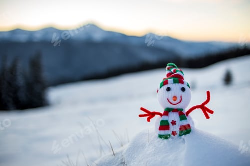 Preview: Small funny toy baby snowman in knitted hat and scarf in deep snow outdoor on blurred snow covered