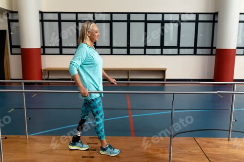 Preview: Senior woman walking in gym using parallel bars with prosthetic leg and running shoes, copy space