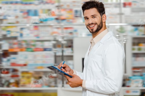 Preview: Selective focus of smiling pharmacist with clipboard looking at camera in apothecary
