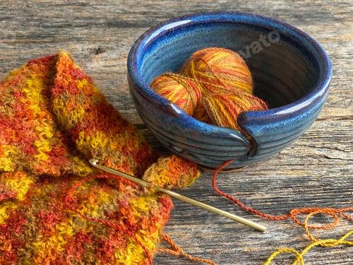 Preview: A blue bowl filled with vibrant balls of yarn on a wooden table along with a crochet hook and scarf!