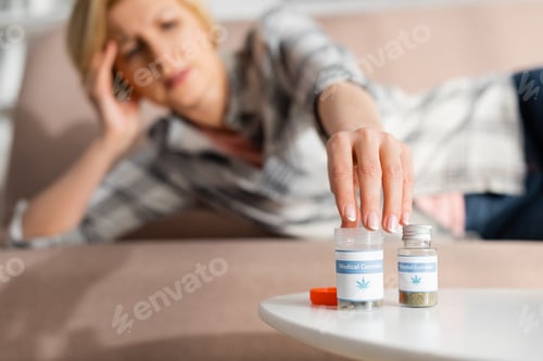 Preview: selective focus of mature woman touching bottle with medical cannabis lettering while lying on sofa