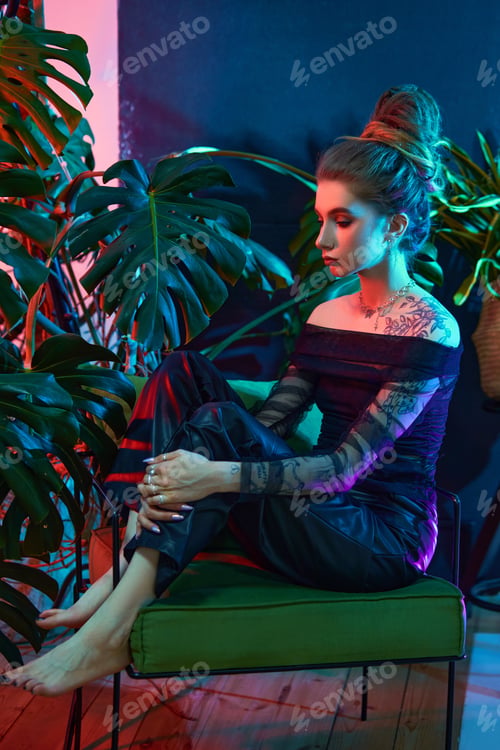Preview: Young woman, wearing a black sheer dress, poses with tropical plants in front of a blue and red