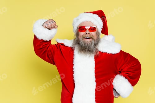 Preview: happy plus size santa claus in trendy sunglasses demonstrating power isolated on yellow