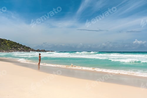 Preview: Woman standing alone on big tropical sandy beach with idyllic turquoise ocean and waves