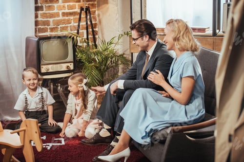 Preview: happy 50s style parents sitting on sofa and looking at adorable children playing at home