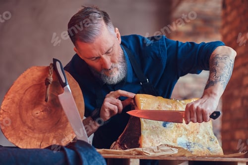 Preview: Bearded elderly butcher cutting huge beefsteak at table