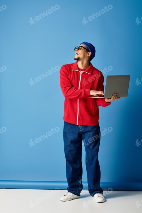 Preview: Creative young man in stylish attire holds laptop against vibrant blue backdrop