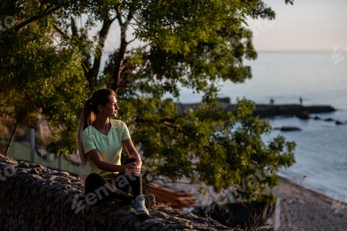 Preview: Fitness athlete runner woman relaxing in sunset. Girl sits on rocks after an active jogging workout.