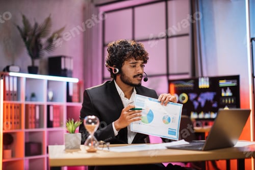 Preview: Young experienced it specialist in headset explaining paper report with graphs, for coworkers