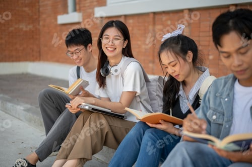 Preview: A positive Asian female student is smiling at the camera, sitting on a footpath with her friends.