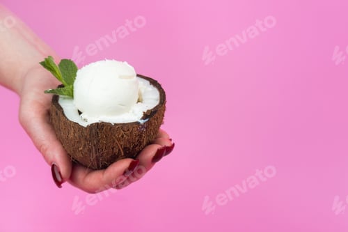 Preview: Vanilla ice cream balls in a fresh coconut half, decorated with mint leaves in your hand on a pink