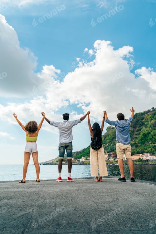 Preview: Vertical shot of diverse friends holding hands with arms raised, celebrating on a pier by the sea.