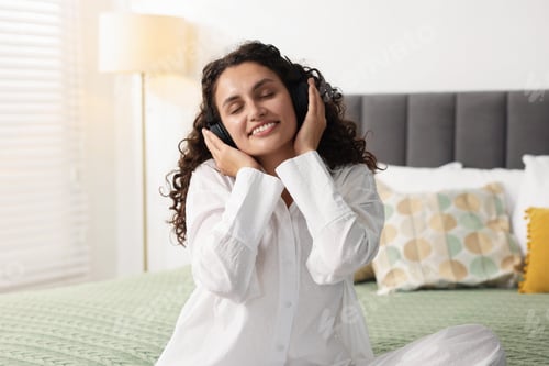 Preview: Young woman with headphones listening to music on bed at home