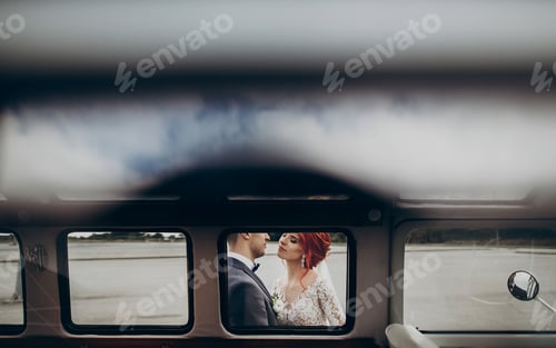 Preview: Stylish groom and happy bride embracing and gently kissing at retro car