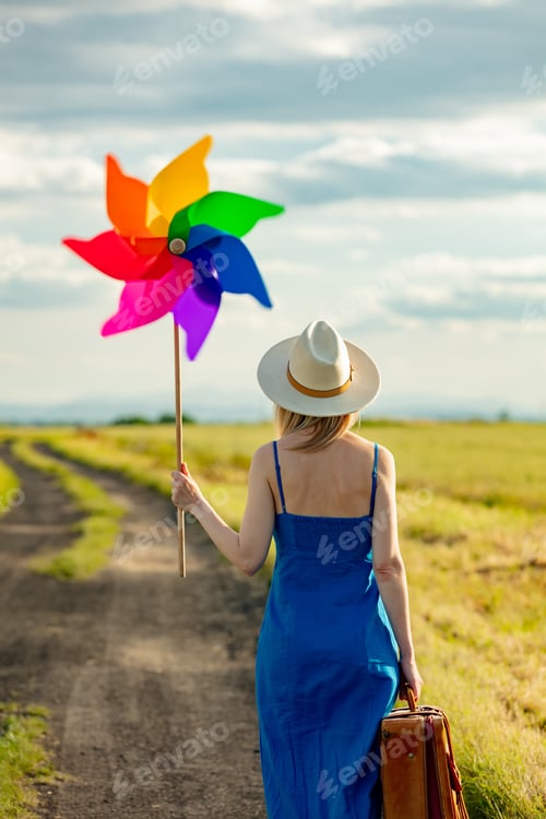 Preview: Girl in blue dress with suitcase and pinwheel on country road in summer.