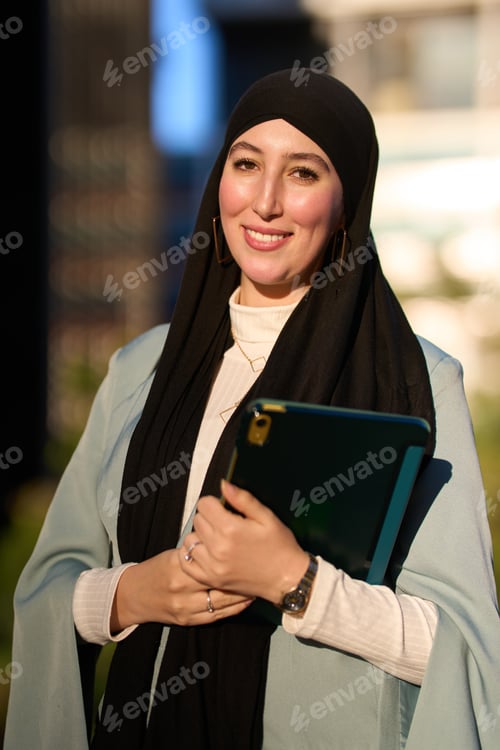 Preview: Smiling muslim businesswoman holding tablet and wearing hijab in urban setting