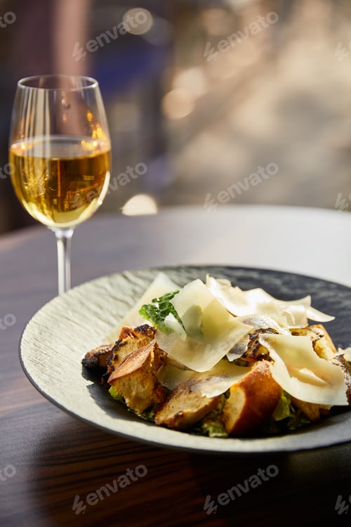 Preview: tasty Caesar salad with Parmesan slices served on plate with white wine in sunlight