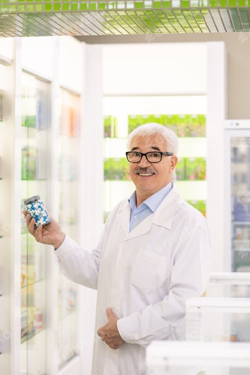 Preview: Aged successful drugstore worker in whitecoat and eyeglasses holding pills