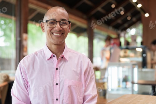 Preview: Happy bald Hispanic man wearing pink casual business shirt and eyeglasses in coffee shop cafe