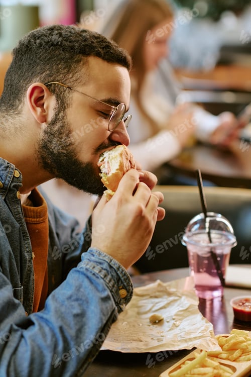 Preview: Young Adult Man Eating Burger at Restaurant Table Indoors