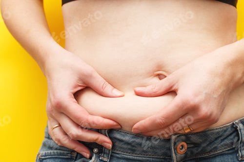 Preview: Woman hands compresses the fat on her belly on a yellow background