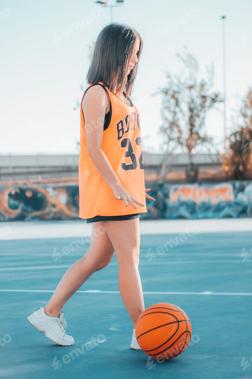 Preview: Vertical shot of an attractive young female in an orange jersey in an outdoor basketball court